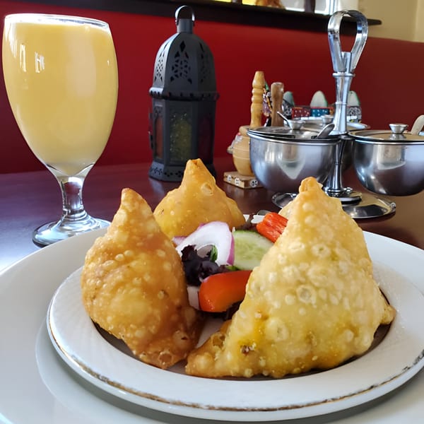 Three golden-fried samosas with a side salad and a glass of mango lassi on a table at Darbar Boston.