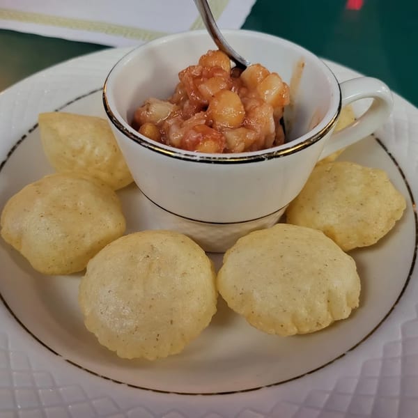 A white cup filled with a chickpea dish, surrounded by six golden-brown puri on a white plate.