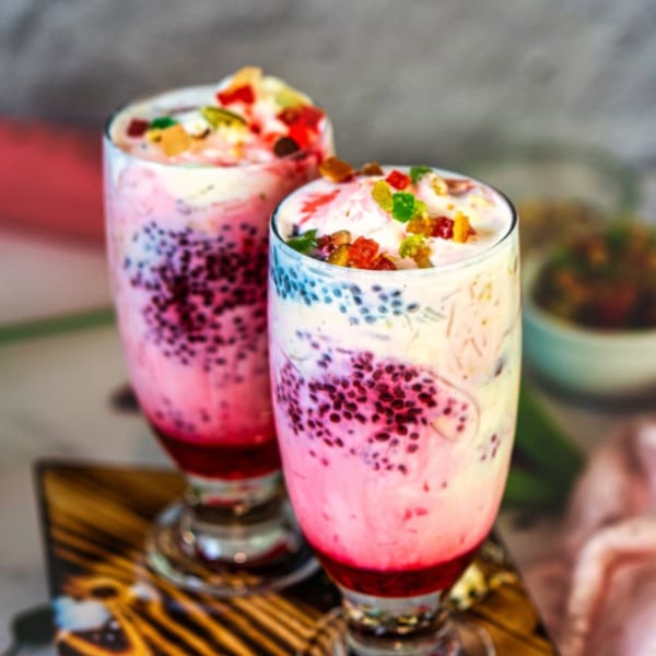 Two glasses of vibrant pink and white falooda, topped with colorful candied fruit, on a wooden tray.